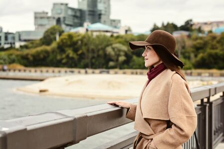 Happy woman in a brown hat and beige coat stands at the edge of the embankment and gazing into the distance. the concept of waiting, of separation, of loveの写真素材