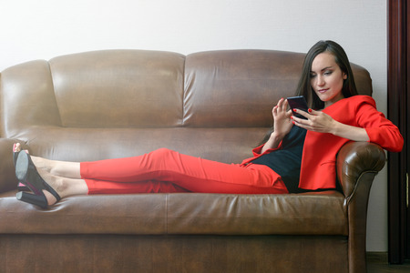 Lazy girl sitting on a leather couch in the office with folded legs and looks to the smartphone. Social networking in the workplace. The concept of idlenessの写真素材
