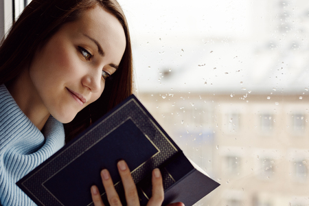 Young brunette woman in blue woolen sweater, reading an interesting book on the windowsill in the room in the rain, cloudy and cold weatherの写真素材