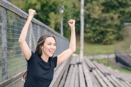 Young brunette woman in a black t-shirt one in the stadium rooting for your favorite team, enjoys raising his hands up and smiles. Emotional expressionの写真素材