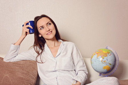 Young smiling woman with a blue Cup of coffee or tea, sitting on the couch in the room and wants to rest, husband, new house, kids, work, travel, cruise, the futureの写真素材