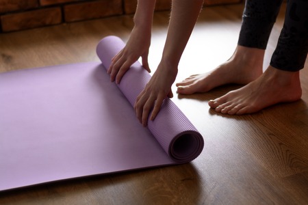 A barefoot woman twists a purple yoga mat and fitness on the parquet floor.の写真素材
