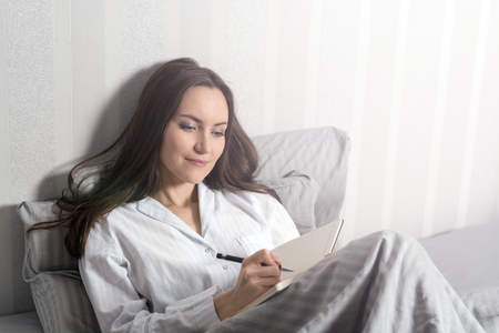Satisfied and happy smiling girl lying on the bed in the room and writes a journal of your dreams, plans, goals, experiences, ideas, lived emotions and feelings.の写真素材