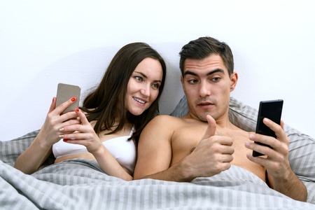 Happy young couple lying in bed and using smartphones. Online shopping, video chatting.の写真素材