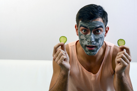 Emotional portrait of a man in a clay mask on his face with cucumbers in his hands. skin care. copy space.の写真素材
