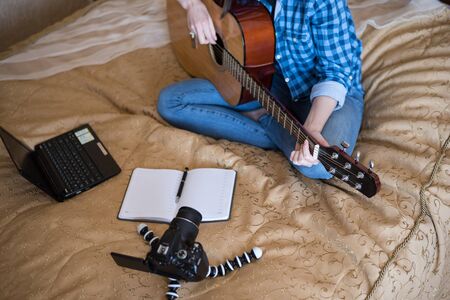 Close - up girl in casual clothes on bed plays acoustic guitar and writes blog on DSLR camera.の写真素材
