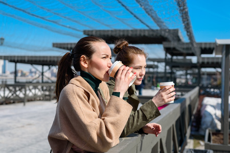 Two women talk on the waterfront with disposable cups of coffee. side view.の写真素材