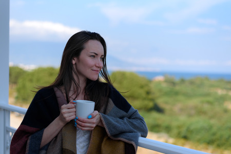 woman in the pochno and a glass of coffee in his hands standing on the balcony of the apartment and...