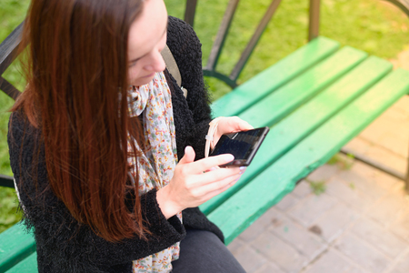 Young brunette sitting on a bench in the Park and uses a smartphone. top view, concept of Internet, social networks, public Wi-Fi.の写真素材