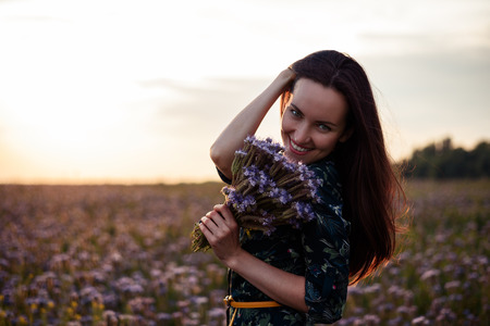 Portrait of a young woman in flower meadow at sunset with a bouquet of purple flowers, smiling, copy spaceの写真素材