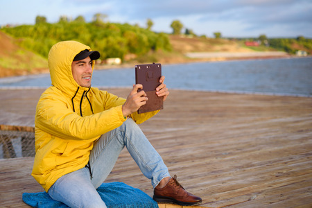 Portrait of man in a yellow raincoat with hood sitting on pier taking photo of sunrise on tablet