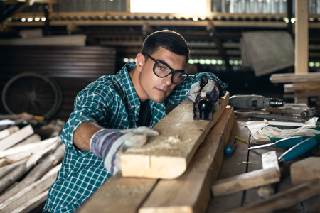Man in plaid shirt, goggles and gloves checks the quality of the planed board in the workshop, manual labor, home craftsmanの写真素材