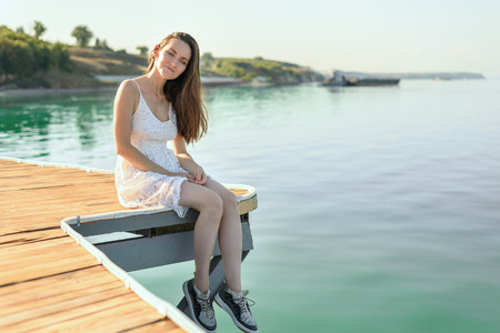 Young relaxed woman in white dress sitting on wooden pier on background of the sea, sunbathing at sunset of dayの写真素材