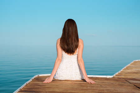 One girl in white dress sitting on pier and looking into distanceの写真素材