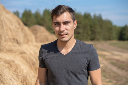 Frontal authentic portrait of young man in gray t-shirt on the background of field with haystacksの写真素材