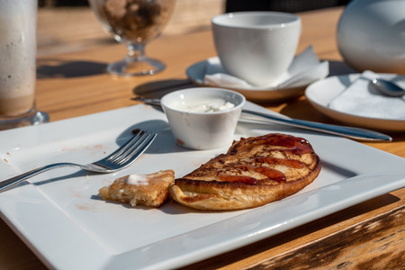 Deadpan image of pancake with honey, sour cream on wooden table with fork, spoon, smartphone and cup...