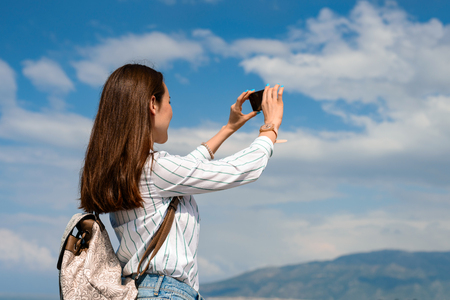 Young woman with backpack outdoors takes a picture of a landscape on a smartphone on the background of blue cloudy sky.の写真素材