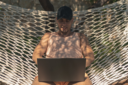 Young guy in a cap, summer clothes sitting in a hammock in a laptop, freelance, 5 g connection, work on vacationの写真素材