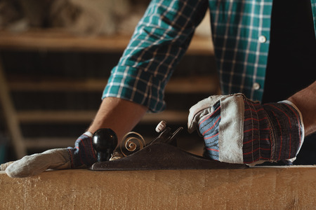 Close-up of men's hands in working gloves with hand plane cut wood. woodworking, roofing, construction, industrial backgroundの写真素材