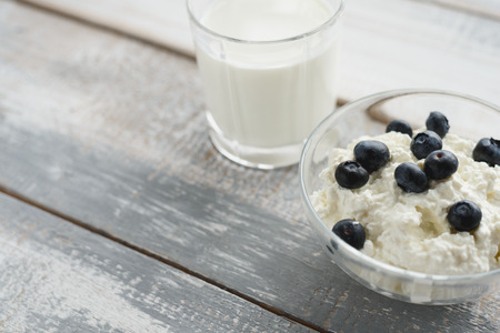 Cottage cheese with blueberries in a glass bowl and a glass of milk on a gray-white wooden background. the concept of a Healthy Breakfast, protein source and calciumの写真素材