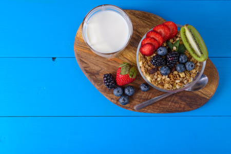 muesli with berries and fruits in glass bowl and glass of milk on blue wooden background. Breakfast...