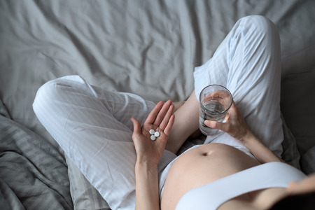 close up of one pregnant woman with pills and glass of water on her hands sitting on the bed. pregnancy conceptの写真素材