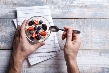 top view oatmeal with berries in glass bowl in the hands of man with spoon on white rustic wooden background. concept of tasty and healthy Breakfastの写真素材