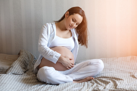 Happy smiling young pregnant woman sitting on bed and holding pregnant tummy. Women's health during pregnancyの写真素材