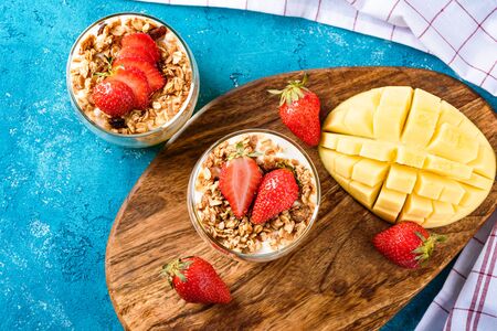 top view, flay lay two glass glasses with mango and strawberry fruit dessert with muesli and cream or ricotta on turquoise backgroundの写真素材