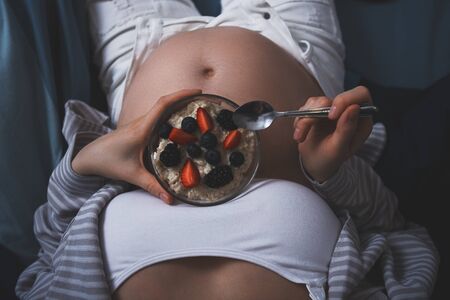 cropped image of pregnant woman in bed with oatmeal with berries, healthy Breakfast in bed, healthy pregnancy, proper healthy dietの写真素材