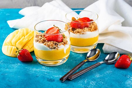 top view two glass glasses with mango and strawberry fruit dessert with muesli and ricotta on turquoise background with dessert spoons and white cotton napkin.の写真素材