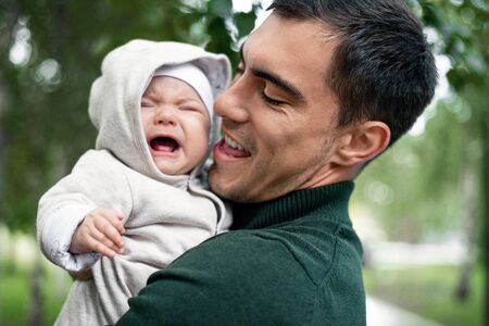 smiling dad in green jacket with crying baby in his arms in the Park