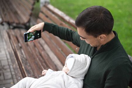 father in green jumper with baby in his arms sitting on bench taking selfie on smartphone