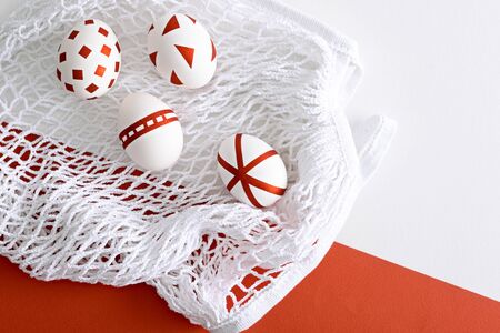 four white Easter eggs with red geometric pattern on white mesh eco bag on red and white background,...