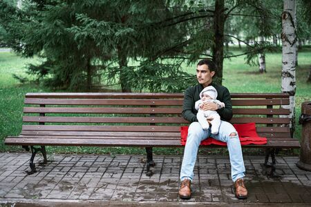 lonely dad in green jacket sitting on bench hugging his baby in Park, fatherhood concept