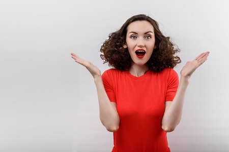 portrait of surprised woman with curly hair in red t-shirt with red lips and raised hands on white background, copy spaceの写真素材