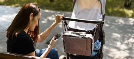 young mother, woman with stroller with smartphone in hands sitting on Park bench, maternity leave...