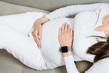top view pregnant woman lying on bed hugging her belly with hands, closeup