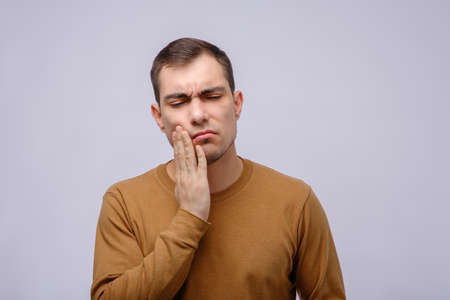 man in beige jacket holds hand to cheek on gray background, toothache, tooth sensitivity, for...