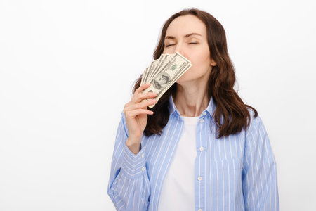 dark-haired woman in blue shirt sniffs dollars that she holds in hand on white background, smell of...