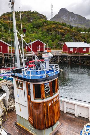 Fishing boat with traditional red wooden houses, rorbuer in the small fishing village of Nusfjord, Lofoten islands, Norway. Nusfjord, Norway - August 26 2019.のeditorial素材