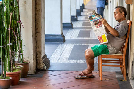 Old man sitting on a chair and reading fresh newspaper. Singapore, Singapore - March 31 2017.のeditorial素材