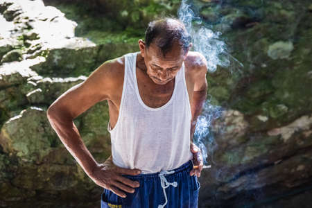 Local old man smoking a cigarette in white t-shirt at Rainforest of Doi Inthanon. Chiang Mai, Thailand - November 10 2017.のeditorial素材