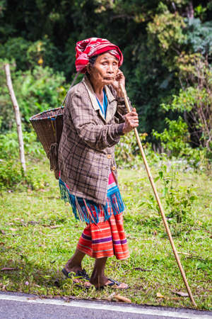 Tribal woman walks by the road in the jungle with a walking stick and talking on a phone. Chiang Mai, Thailand - November 11 2017.のeditorial素材