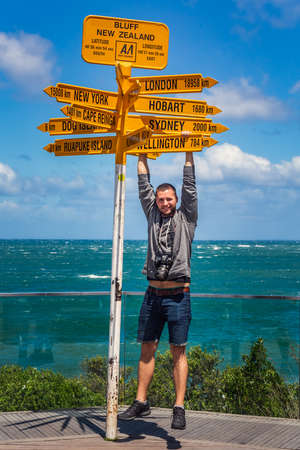 Man hangs from the world's southernmost signpost in Bluff, South Island. Global signpost shows world distances measured from Bluff, tourist destination. Bluff, New Zealand - December 19 2017.のeditorial素材