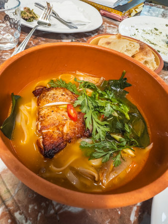 A rich and flavorful spicy chicken noodle soup served in a clay bowl, topped with fresh herbs like...