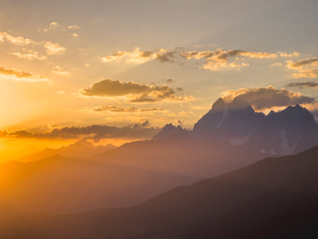 A mesmerizing sunset view over the Caucasus Mountains, casting warm hues and silhouettes across the peaks. Soft clouds scatter the sky, enhancing the golden glow and creating a serene landscape.の写真素材