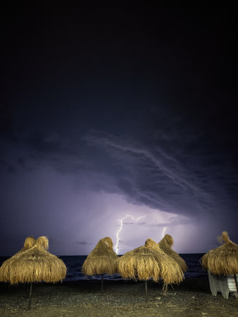 Dramatic lightning illuminates a dark sky above a beach lined with straw umbrellas, creating a striking contrast between natures power and seaside tranquility.の写真素材