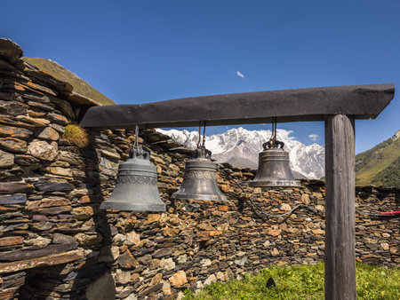 Traditional metal bells hang against a backdrop of a stone wall and majestic snow-covered mountains in Svaneti, Georgia, offering a stunning view of the Caucasus.の写真素材
