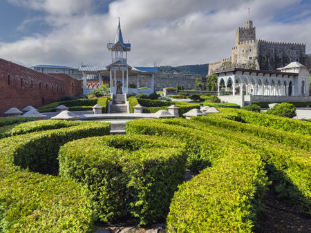 A picturesque garden with sculpted hedges, a pavilion, and a historic fortress with arched colonnades and stone walls in the background.の写真素材